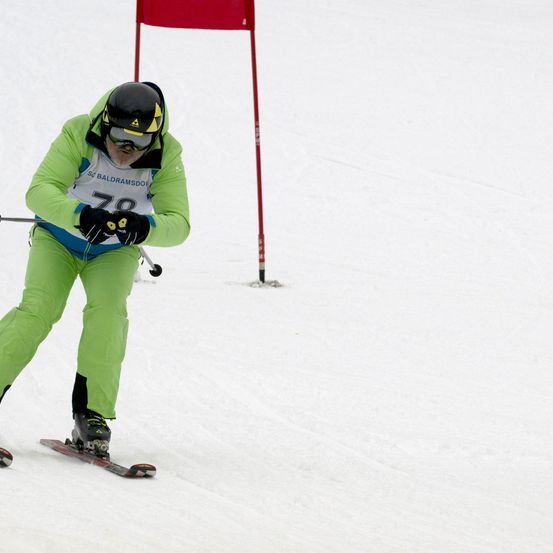 Ein Skifahrer in einem grünen Skianzug mit der Nummer 78 fährt einen schneebedeckten Hang hinunter. Hinter ihm steht eine rote Flagge.