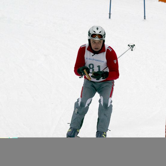 Ein Skifahrer mit der Nummer 81 auf seinem roten und weißen Trikot fährt einen verschneiten Hang hinunter. Er trägt einen weißen Helm, eine Brille und Handschuhe.