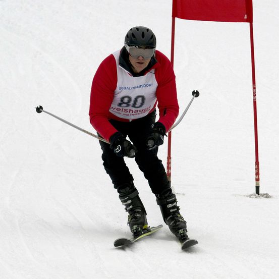 Ein Skifahrer, der ein rotes und weißes Langarmshirt mit der Nummer 80 trägt, fährt einen Schneehang hinunter, hält Skistöcke in der Hand, mit einer roten Fahne im Hintergrund.