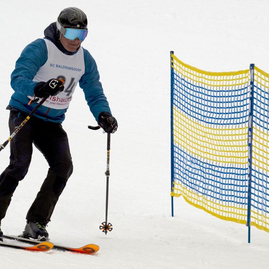 Ein Skifahrer mit blauer Jacke und Nummer 4 fährt einen verschneiten Hang hinunter, mit einem gelben und blauen Netzzaun.