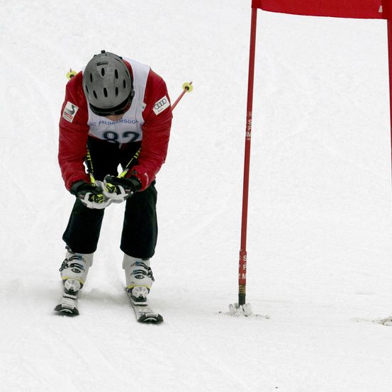 Ein Skifahrer, der einen roten Anzug, schwarze Hosen und einen Helm trägt, navigiert durch einen verschneiten Parcours und berührt leicht eine rote Stange.