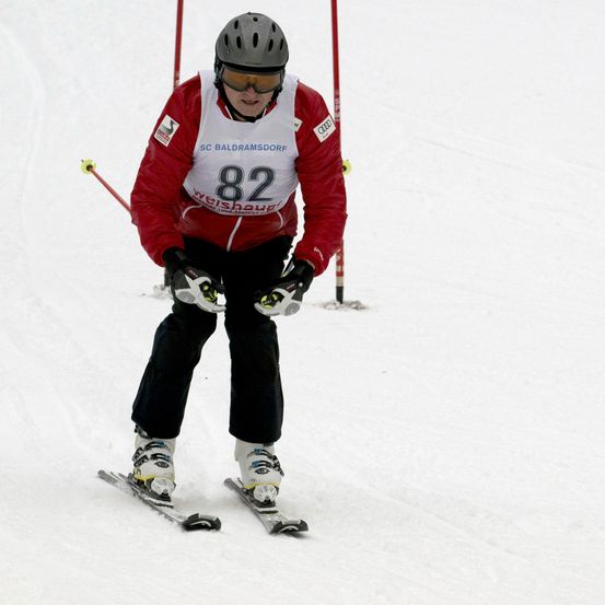 Ein Skifahrer in einem roten Jacke, schwarzen Hosen und einem grauen Helm skiiert auf einem verschneiten Hang. Die Nummer 82 ist auf der Vorderseite seiner Jacke angebracht.