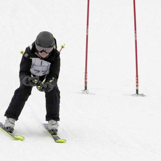 Ein Skifahrer mit Helm und Schutzbrille fährt einen schneebedeckten Hang hinunter und navigiert um rote Pfosten herum.