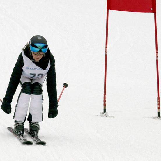 Ein Skifahrer mit Helm, Schutzbrille und weißer Jacke mit SC Baldramslev. Sie fahren einen verschneiten Hang hinunter und navigieren um rote Flaggen herum.