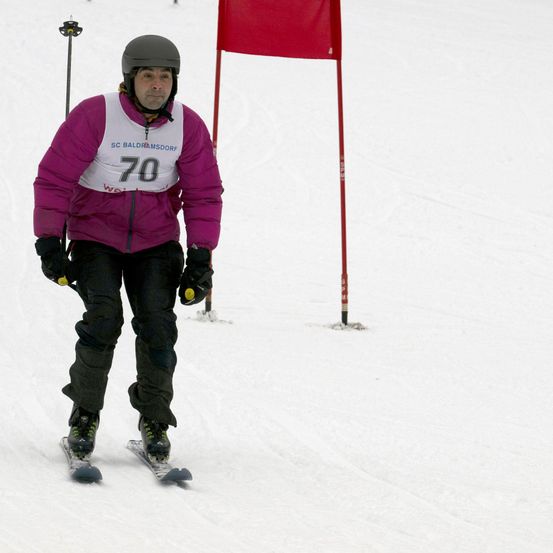 Ein Skifahrer mit der Nummer 70 auf seiner Jacke fährt einen schneebedeckten Hang hinunter, trägt Skiausrüstung und hält Skistöcke. Hinter ihm steht eine rote Fahne.