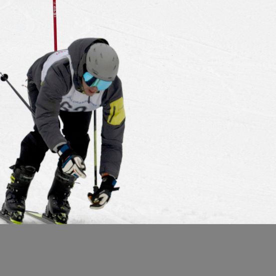 Ein Skifahrer bewegt sich auf einer verschneiten Piste, trägt einen Helm, eine Brille und Skiausrüstung. Der Skifahrer hat die Nummer 62 auf seiner Jacke. Der Skifahrer hält Skistöcke in der Hand.