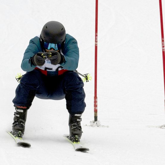 Ein Skifahrer in einer blauen Jacke und schwarzem Helm fährt einen verschneiten Hang hinunter und navigiert zwischen zwei roten Pfosten.