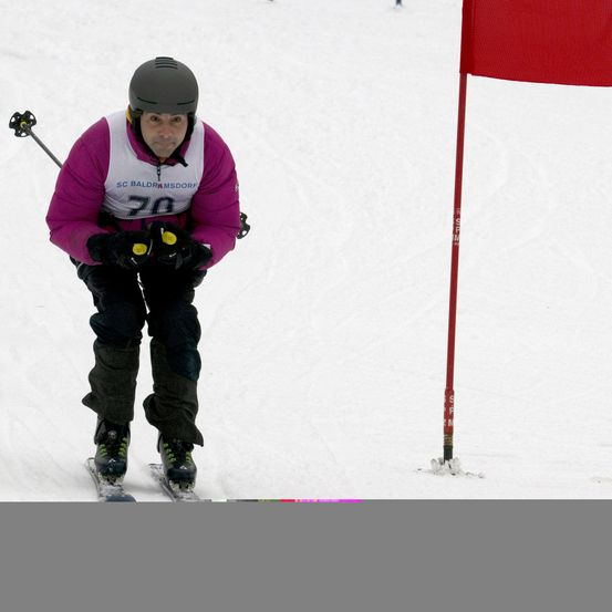 Ein Skifahrer in einem lilafarbenen Jacke mit der Nummer 70 fährt einen schneebedeckten Hang hinunter und hält Skistöcke. Ein roter Flagge steht im Hintergrund.