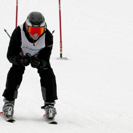 Ein Skifahrer fährt bergab auf einer verschneiten Piste. Er trägt einen Helm, eine Brille und einen Skianzug mit dem Text 'SC BA DRAMSD'.