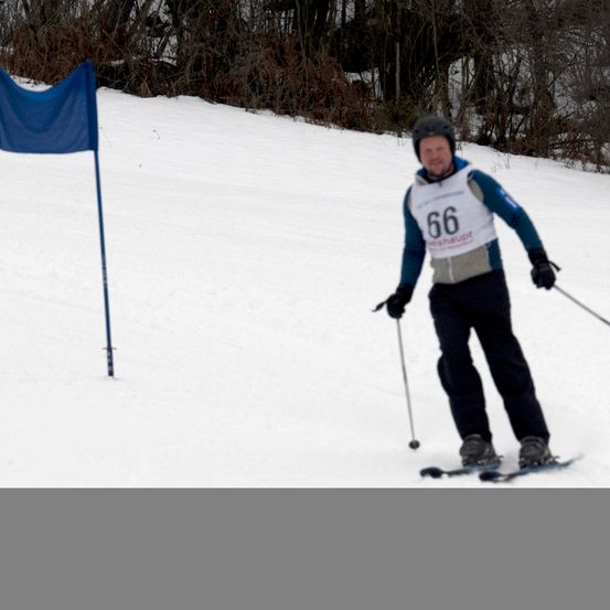 Ein Skifahrer mit der Nummer 66 navigiert einen verschneiten Kurs und hält Skistöcke. Eine blaue Fahne ist links neben dem Weg positioniert.