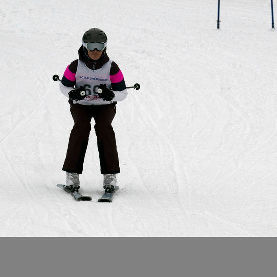 Ein Skifahrer fährt einen verschneiten Hang hinunter, trägt einen Helm und eine Schutzbrille. Sie halten Skistöcke in den Händen und ihre Startnummer ist 66.