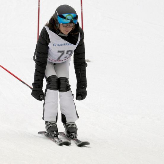 Ein Skifahrer in einem schwarzen Helm und Schutzbrille, trägt ein schwarz-weißes Trikot mit 'SC BALDRAMSD' und '73', skifahrer auf Schnee.