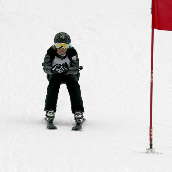 Ein Skifahrer navigiert durch eine verschneite Piste und passiert eine rote Flagge. Er trägt einen Helm, eine Brille und Handschuhe.