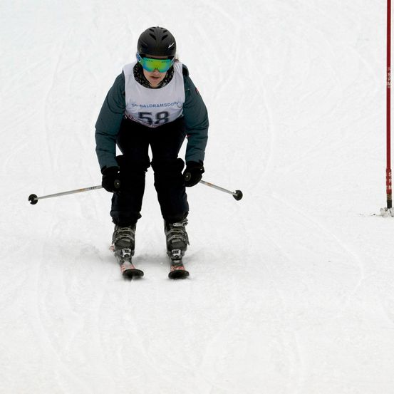 Ein Skifahrer mit Helm, Brille und Handschuhen fährt auf einer verschneiten Piste. Er hält Skistöcke in der Hand und scheint in Bewegung zu sein.