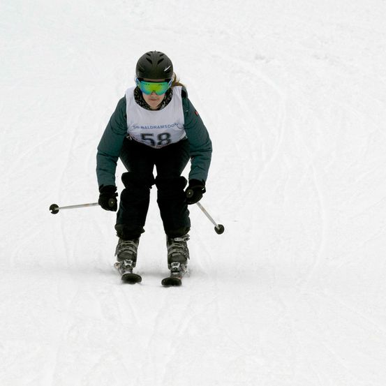 Ein Skifahrer mit der Nummer 58 auf seiner Jacke fährt einen schneebedeckten Hang hinunter, trägt einen Helm und eine Schutzbrille.