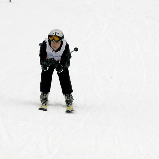 Ein Skifahrer in einer weißen Jacke mit Brille und Handschuhen fährt einen verschneiten Hang hinunter und hält einen Skistock in jeder Hand.