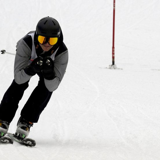 Ein Skifahrer mit schwarzem Helm, Sonnenbrille und Handschuhen fährt einen Schneehang hinunter.