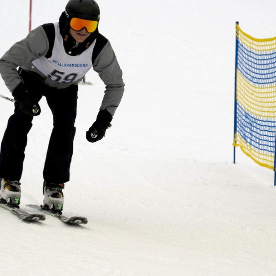 Ein Skifahrer mit der Nummer 59 auf dem Brustanhänger fährt auf einer verschneiten Piste. Er trägt einen Helm, eine Brille und Skiausrüstung. Eine gelbe und blaue Netzabsperrung befindet sich an der Seite.