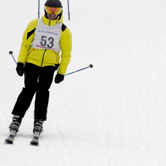 Ein Skifahrer in einem gelben Jackett mit der Nummer 53, einer weißen Nummer, einer schwarzen Brille und schwarzen Handschuhen fährt einen schneebedeckten Hang hinunter.
