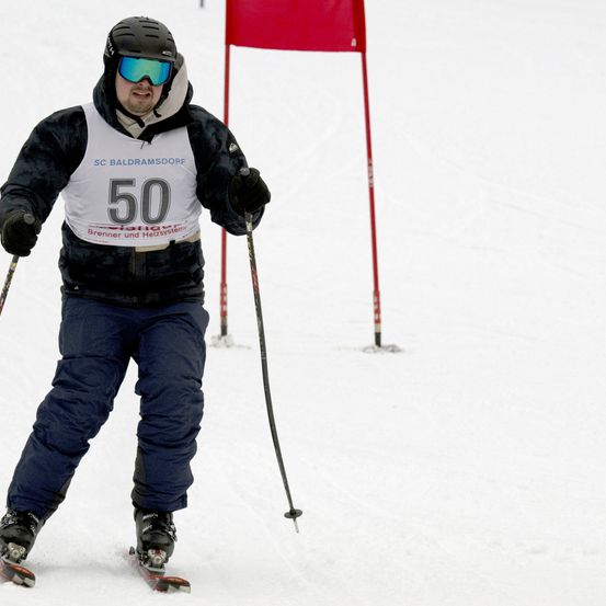 Ein Skifahrer mit der Nummer 50 auf seinem Bib ist auf einem verschneiten Hang unterwegs, hinter ihm steht eine rote Fahne.