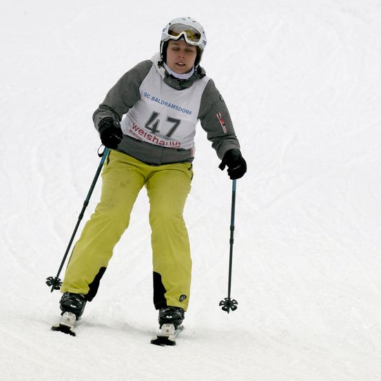 Ein Skifahrer mit der Nummer 47 auf der Jacke fährt einen verschneiten Hang hinunter, trägt eine Skibrille und Skischuhe.