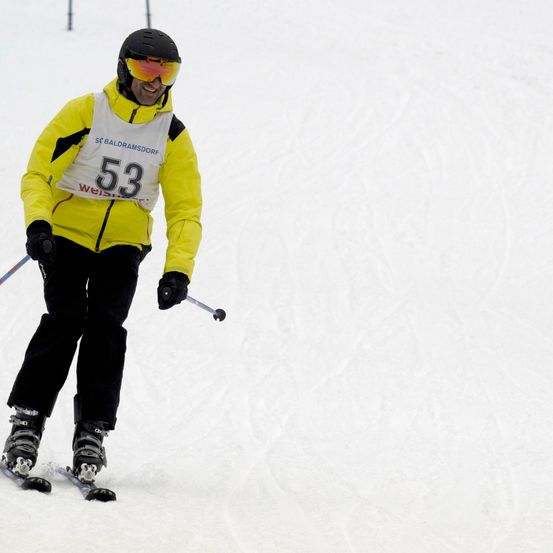 Ein Skifahrer mit der Nummer 53 auf seiner Jacke fährt einen schneebedeckten Hang hinunter. Er trägt einen Helm und eine Schutzbrille.