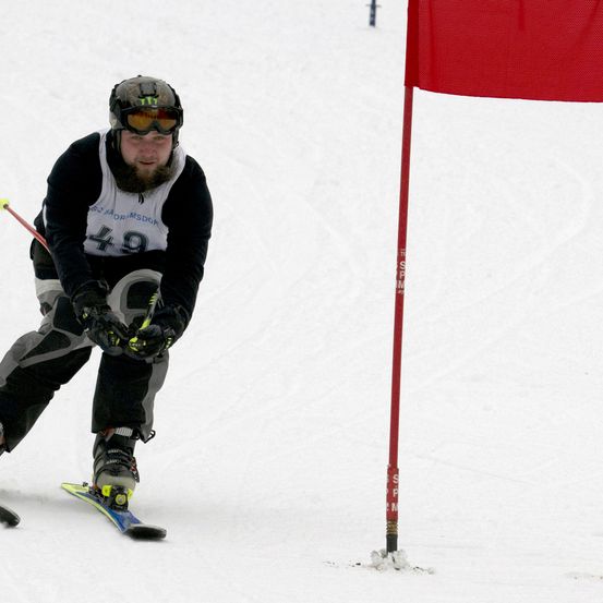 Ein Skifahrer mit der Nummer 49 auf seinem Startnummernband navigiert eine Piste und passiert einen roten Flaggenpfosten. Er trägt einen Helm, eine Skibrille und Skiausrüstung.