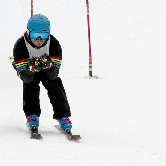 Ein Skifahrer mit blauem Helm, Schutzbrille und bunter Skiausrüstung fährt einen schneebedeckten Hang hinunter. Er beugt sich leicht nach vorne, möglicherweise um das Gleichgewicht zu halten. Rote Stangen sind im Hintergrund zu sehen.