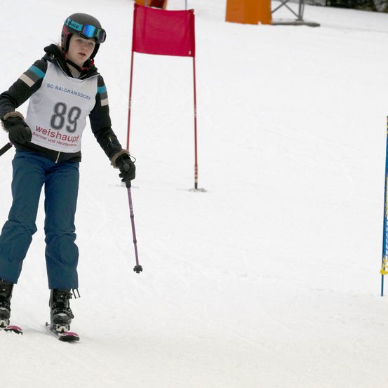 Ein Skifahrer mit Helm, Schutzbrille und einem weißen Trikot mit der Nummer 89, der einen verschneiten Hang hinunterfährt. Dahinter steht eine rote Fahne.