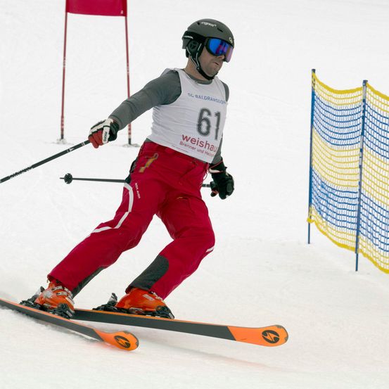 Ein Skifahrer in roten Skihosen und einem weißen Trikot mit der Nummer 61 fährt bergab. Er trägt einen schwarzen Helm, Handschuhe und eine Schutzbrille.