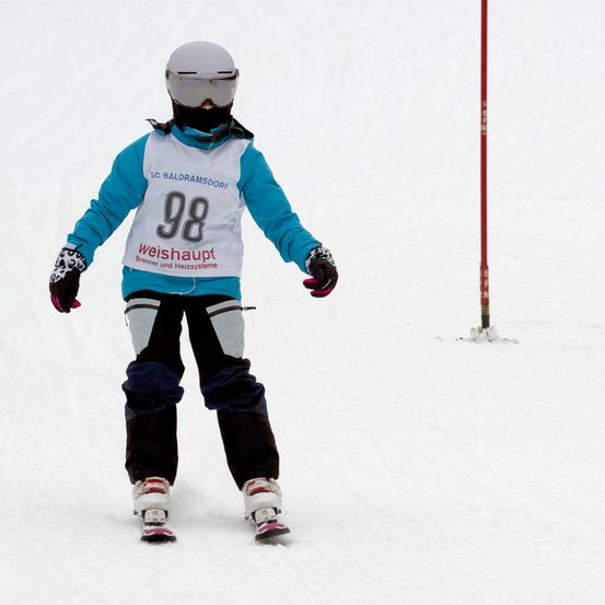 Ein Skifahrer mit weißem Helm und Schutzbrille in einer blau-weißen Skijacke mit der Nummer 98 und dem Wort weishaupt fährt einen schneebedeckten Hang hinunter.