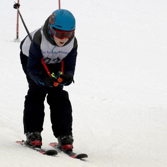 Ein junger Skifahrer fährt einen schneebedeckten Hang hinunter, trägt einen blauen Helm, eine Brille und Skiausrüstung. Der Skifahrer hält einen Skistock in der rechten Hand.