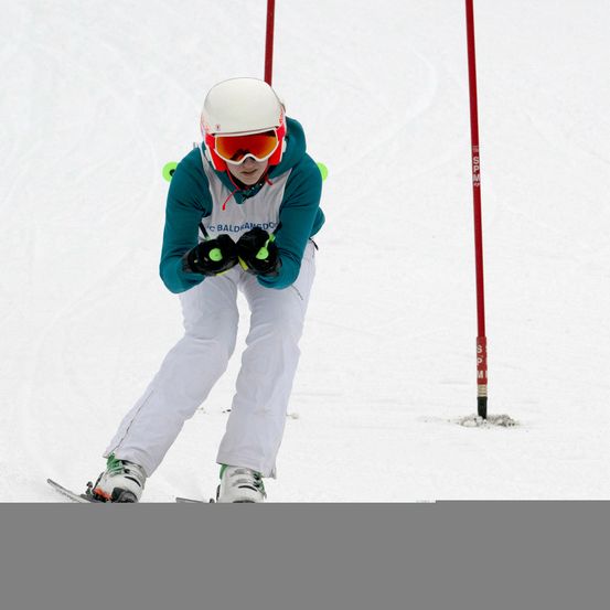 Ein Skifahrer in weißer Ausrüstung und mit Schneebrille fährt auf einem verschneiten Hang, mit Pfosten, die den Kurs markieren.