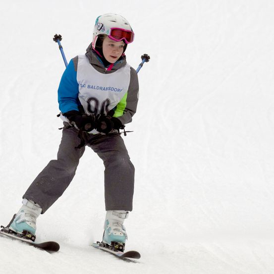 Ein junger Skifahrer mit Helm und Schutzbrille fährt bergab. Auf der Jacke steht die Nummer '99' und der Skifahrer hält Skistöcke in den Händen.