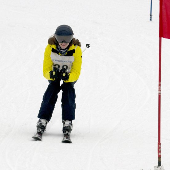 Ein Skifahrer fährt bergab auf einem verschneiten Hang, trägt eine gelbe Jacke mit der Nummer 38. Der Skifahrer hält einen Skistock in beiden Händen. Der Skifahrer trägt Helm und eine Schutzbrille. Eine rote Flagge befindet sich auf der rechten Seite des Bildes.