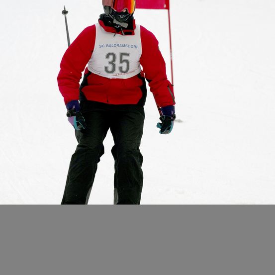 Ein Skifahrer mit der Nummer 35 auf seiner Jacke fährt einen verschneiten Hang hinunter. Er trägt eine Skibrille, einen Helm und Handschuhe. Hinter ihm steht eine rote Fahne.