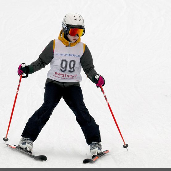 Ein Skifahrer in einer weißen und grauen Jacke mit der Nummer 99 fährt einen schneebedeckten Hang hinunter, trägt eine Schutzbrille und hält Skistöcke in den Händen.