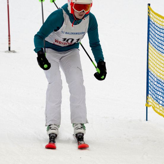 Ein Skifahrer in weißer Schneekleidung fährt einen Schneehang hinunter. Er hält Skistöcke und trägt eine Schneebrille.