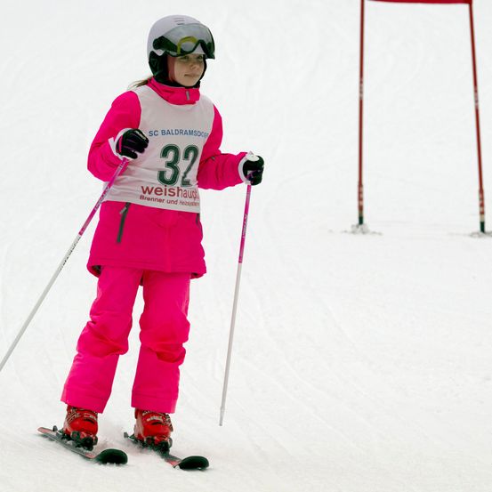 Eine junge Skifahrerin in pinker Ausrüstung navigiert einen schneebedeckten Hang. Sie trägt ein weißes Startnummernband mit der Nummer 32 und hält Skistöcke.