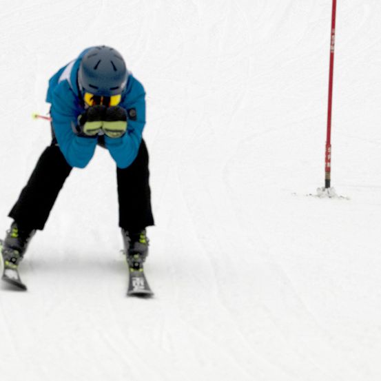 Ein Skifahrer in einem blauen Jackett und Helm fährt auf einem verschneiten Hang und verfehlt knapp einen roten Pfosten.