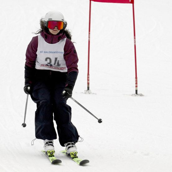 Ein junger Skifahrer mit weißem Helm und Schutzbrille fährt bergab. Er trägt eine Nummer 34 und einen bordeauxfarbenen Mantel. Dahinter stehen rote Stangen mit einer rosa Flagge.