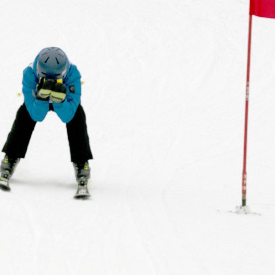 Ein Skifahrer in blauer Jacke und Helm steht auf schneebedecktem Boden, gebeugt, als ob er sich zum Skifahren bereit macht, mit einer roten Fahne im Hintergrund.