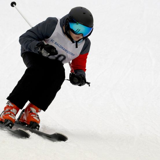 Ein junger Skifahrer mit schwarzem Helm und Schutzbrille fährt einen schneebedeckten Hang hinunter. Die Skier und Skischuhe des Skifahrers sind orange.