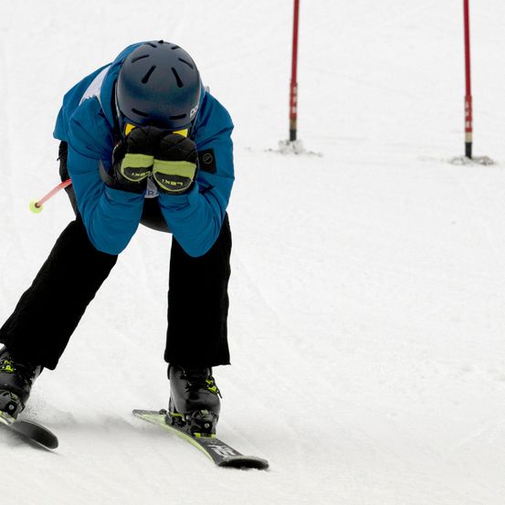 Ein Skifahrer in einer blauen Jacke, schwarzen Hosen und Skiausrüstung fährt den Schneehang hinunter und hält Skistöcke.