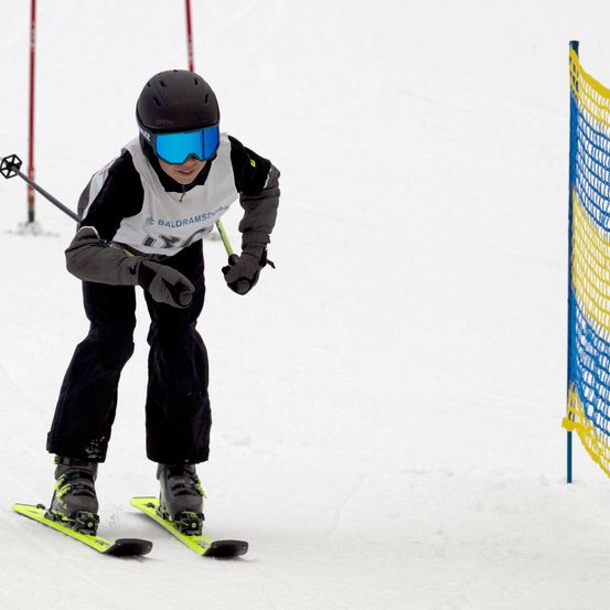 Ein Skifahrer mit schwarzem Helm und blauen Skibrille fährt den Schneehang hinunter. Er hält Skistöcke und trägt einen weißen und schwarzen Skianzug. Rechts ist ein blaues und gelbes Netz zu sehen.