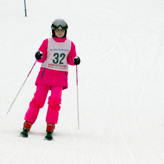 Ein junger Skifahrer in einem rosa Skianzug mit der Nummer 32 fährt auf einer verschneiten Piste. Der Skifahrer hält Skistöcke und trägt eine Skibrille und Handschuhe.