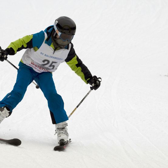 Ein Skifahrer mit schwarzem Helm und Schutzbrille fährt einen schneebedeckten Hang hinunter. Er trägt einen blau-grünen Skianzug mit der Nummer 25.