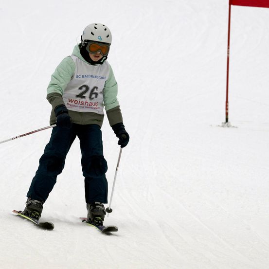 Ein Skifahrer mit weißem Helm und Schutzbrille fährt einen schneebedeckten Hang hinunter, im Hintergrund ein roter Flaggenpfosten.