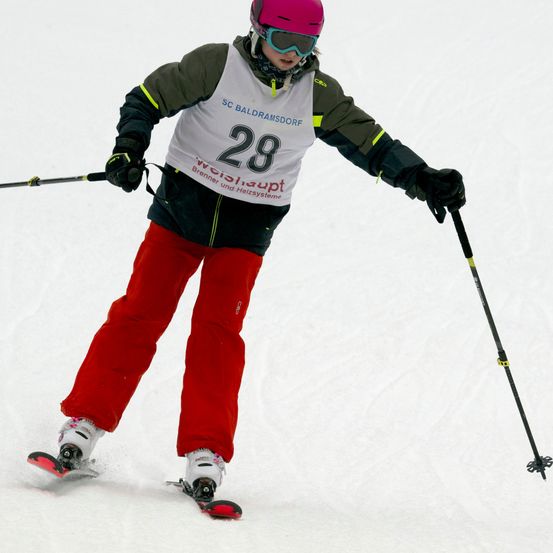 Ein Skifahrer mit einem Trikot der Nummer 28 fährt einen verschneiten Hang hinunter. Sie tragen einen rosa Helm und eine Schutzbrille.