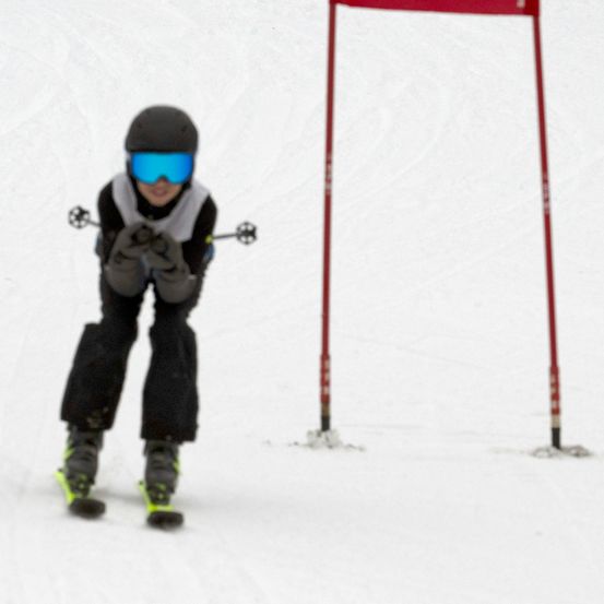 Ein Skifahrer fährt bergab und trägt schwarze Skihosen, eine schwarze Skibrille und einen schwarzen Helm. Im Hintergrund befindet sich eine rote Flagge.